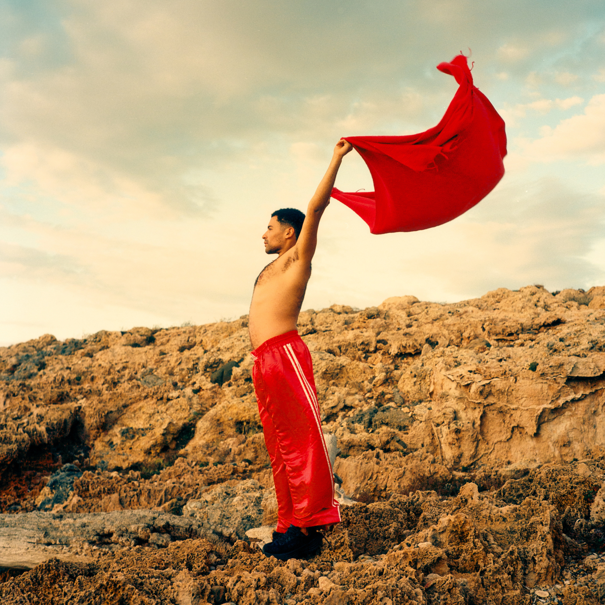 Man in red pants holding a red fouta against a rocky landscape with a cloudy sky.
