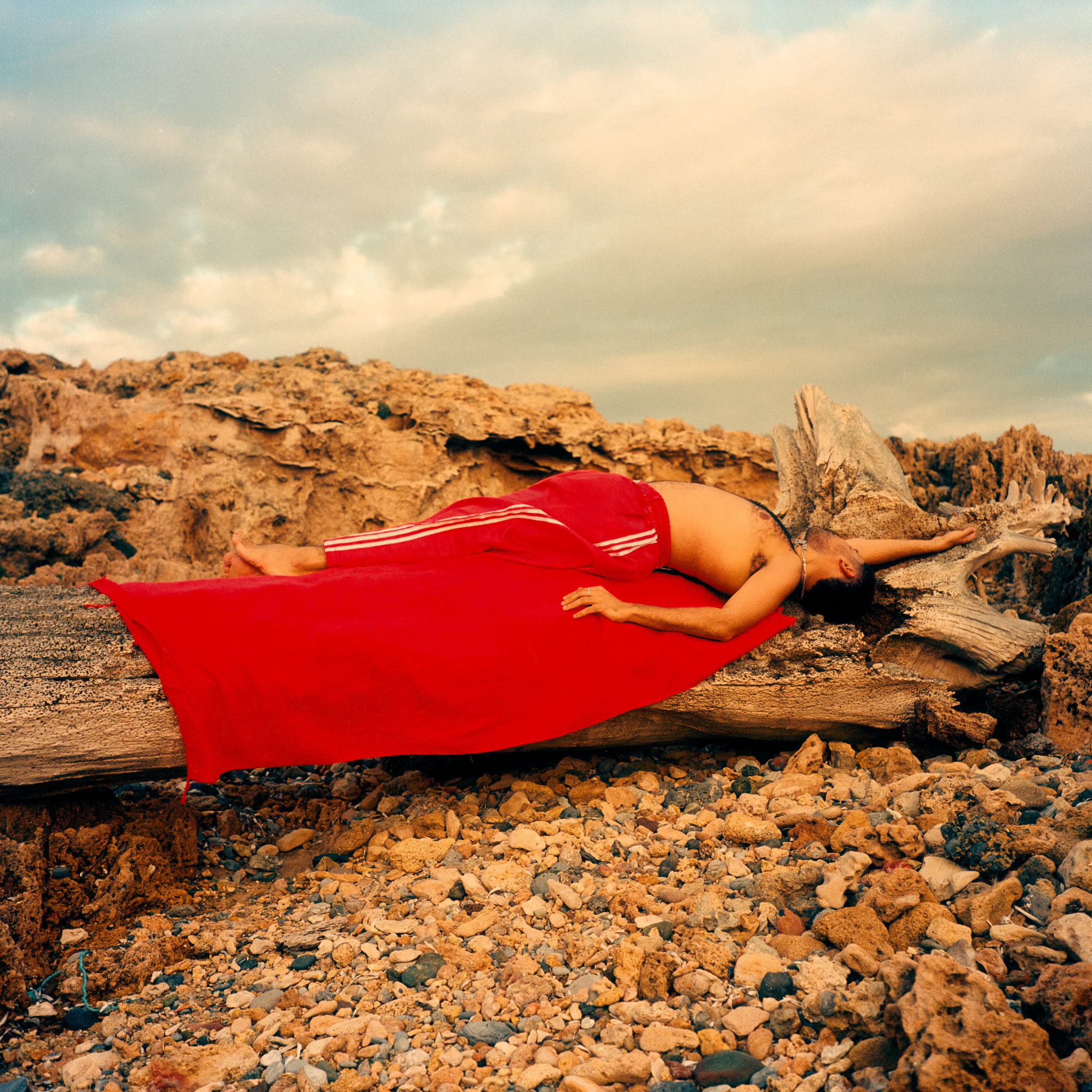 Person lying on a red fouta in a rocky landscape with a cloudy sky.