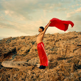 Man holding a red fouta above his head on a rocky landscape with a cloudy sky.