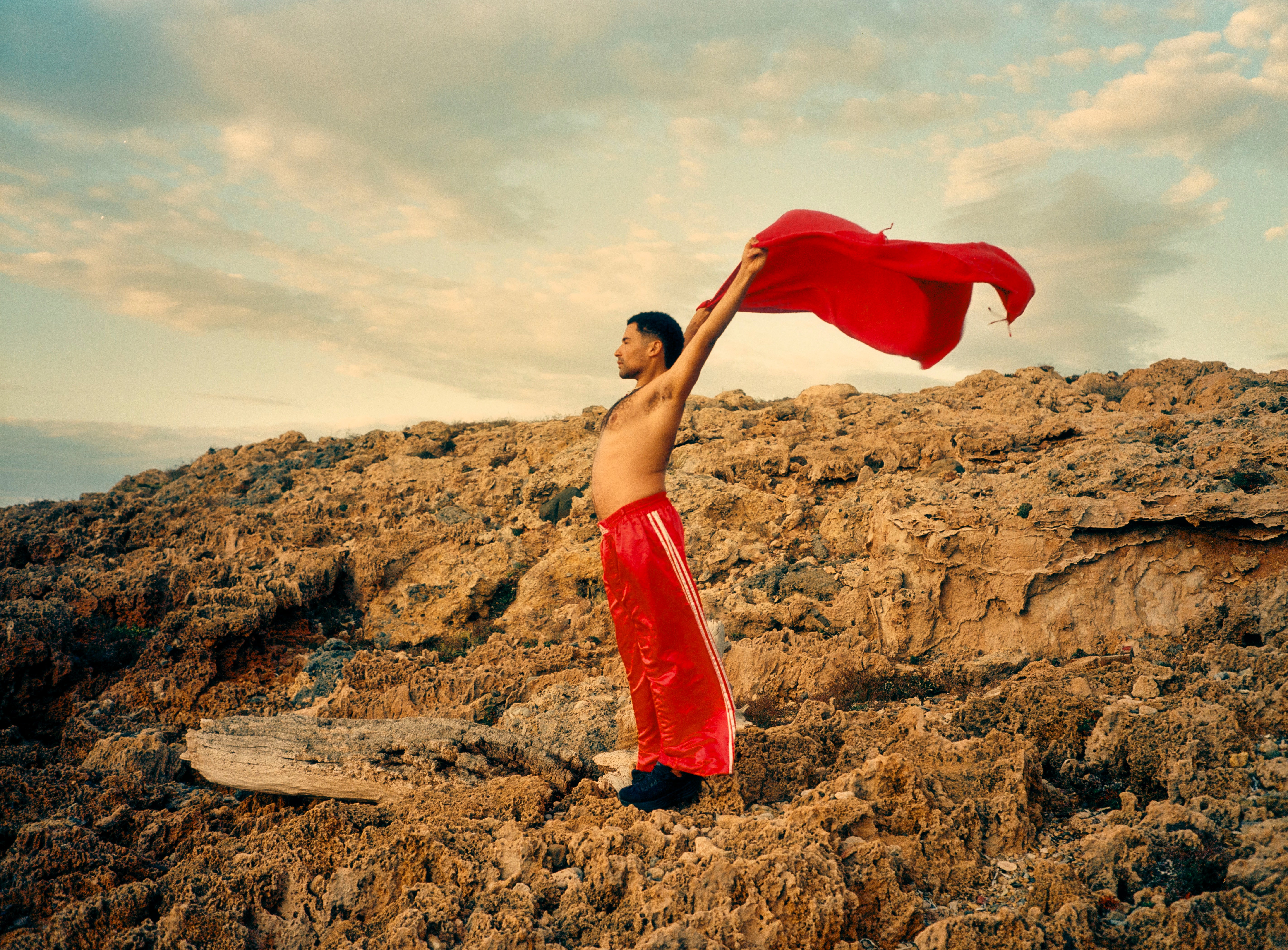 Man holding a red fouta above his head on a rocky landscape with a cloudy sky.